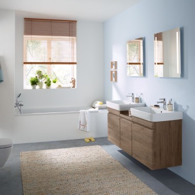 Rodinná kúpeľňa so svetlomodrou stenou a kúpeľňovým nábytkom v drevenom dizajne hickory, zrkadlovou skrinkou, ovládacím tlačidlom a sanitárnou keramikou od spoločnosti Geberit.